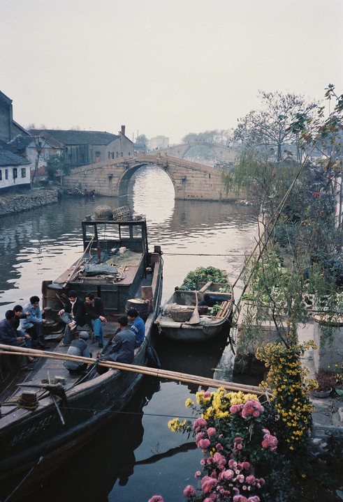 Several men are gathered on a boat in a canal with an arched stone bridge in the background and colorful flowers in the foreground.