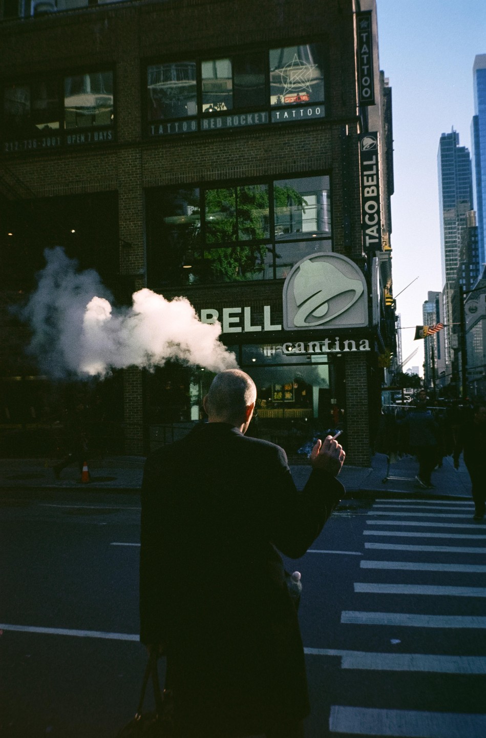 A man stands on a street corner, exhaling a large cloud of smoke, with a Taco Bell sign and a tattoo parlor in the background.