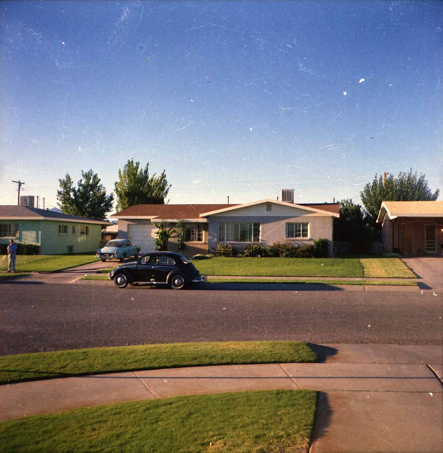 A man stands on a lawn next to a street with two cars parked in front of houses.