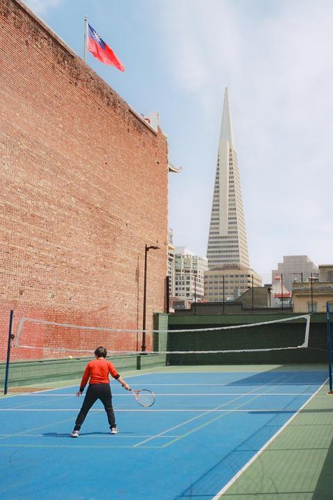 A person stands on a blue tennis court with a racket, facing a net, with a tall building and a flag visible in the background.