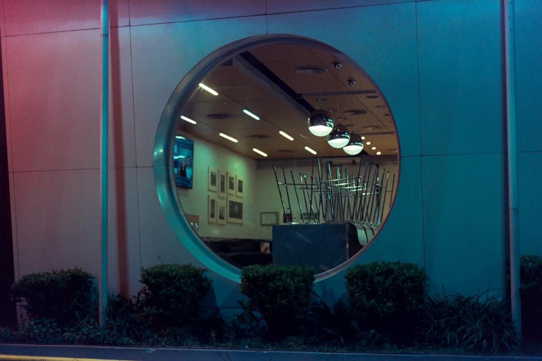 A round window reveals the interior of a closed restaurant with chairs stacked on tables, illuminated by neon light.