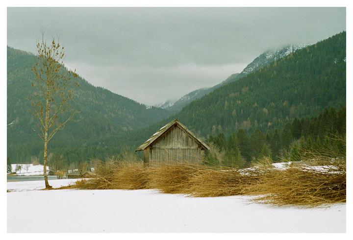 A wooden barn sits in a snowy field with mountains and a forest in the background.