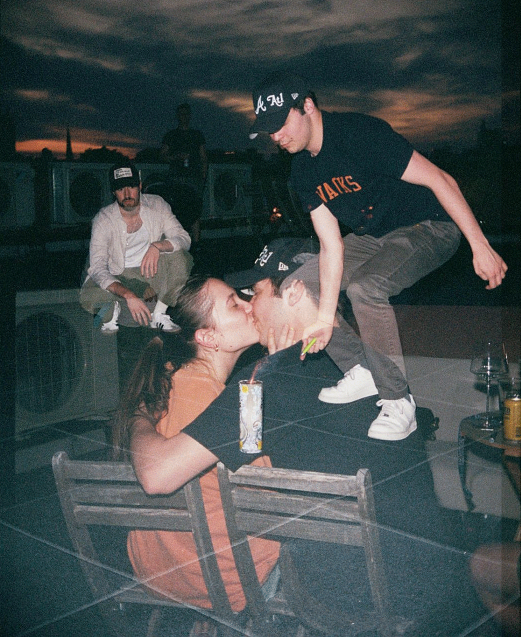 A couple kisses on a rooftop at dusk while other people are visible in the background.
