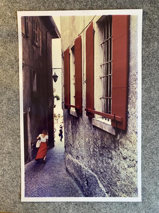 A woman in a red skirt walks down a narrow cobblestone alleyway between old buildings with red shutters.