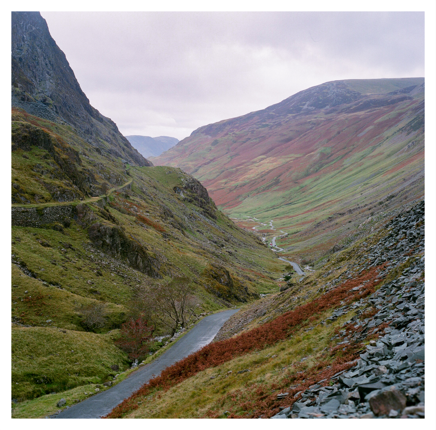 A winding road and a river run through a valley with steep, rocky, and grassy slopes under a cloudy sky.