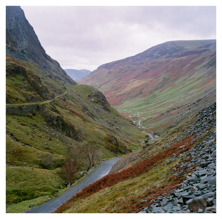 A winding road and a river run through a valley with steep, rocky, and grassy slopes under a cloudy sky.