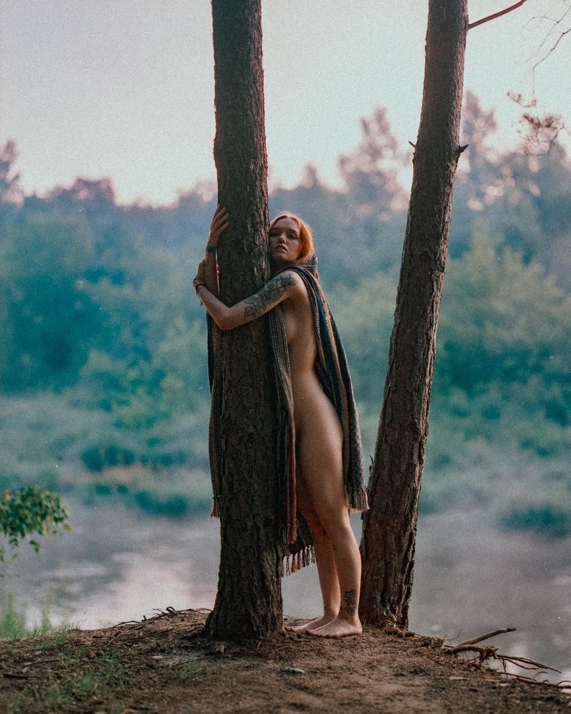 A woman wrapped in a blanket stands between two trees on a hill overlooking a body of water.
