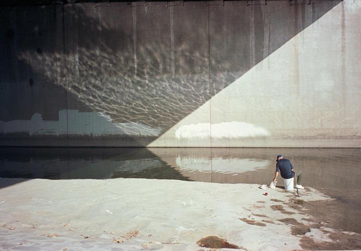 A man is fishing in a concrete river with light reflecting off the water onto the wall.