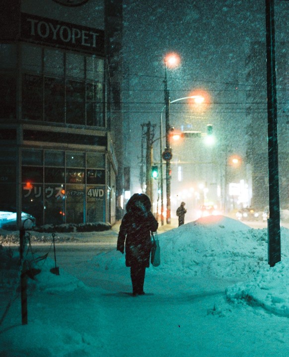 A person walks down a snowy street at night, past a Toyopet dealership and streetlights.