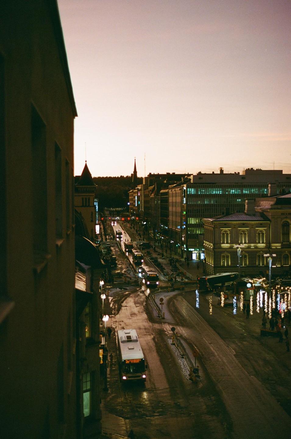 A street with buses and cars is viewed from above at dusk, with buildings lining both sides of the road and a light-colored sky overhead.