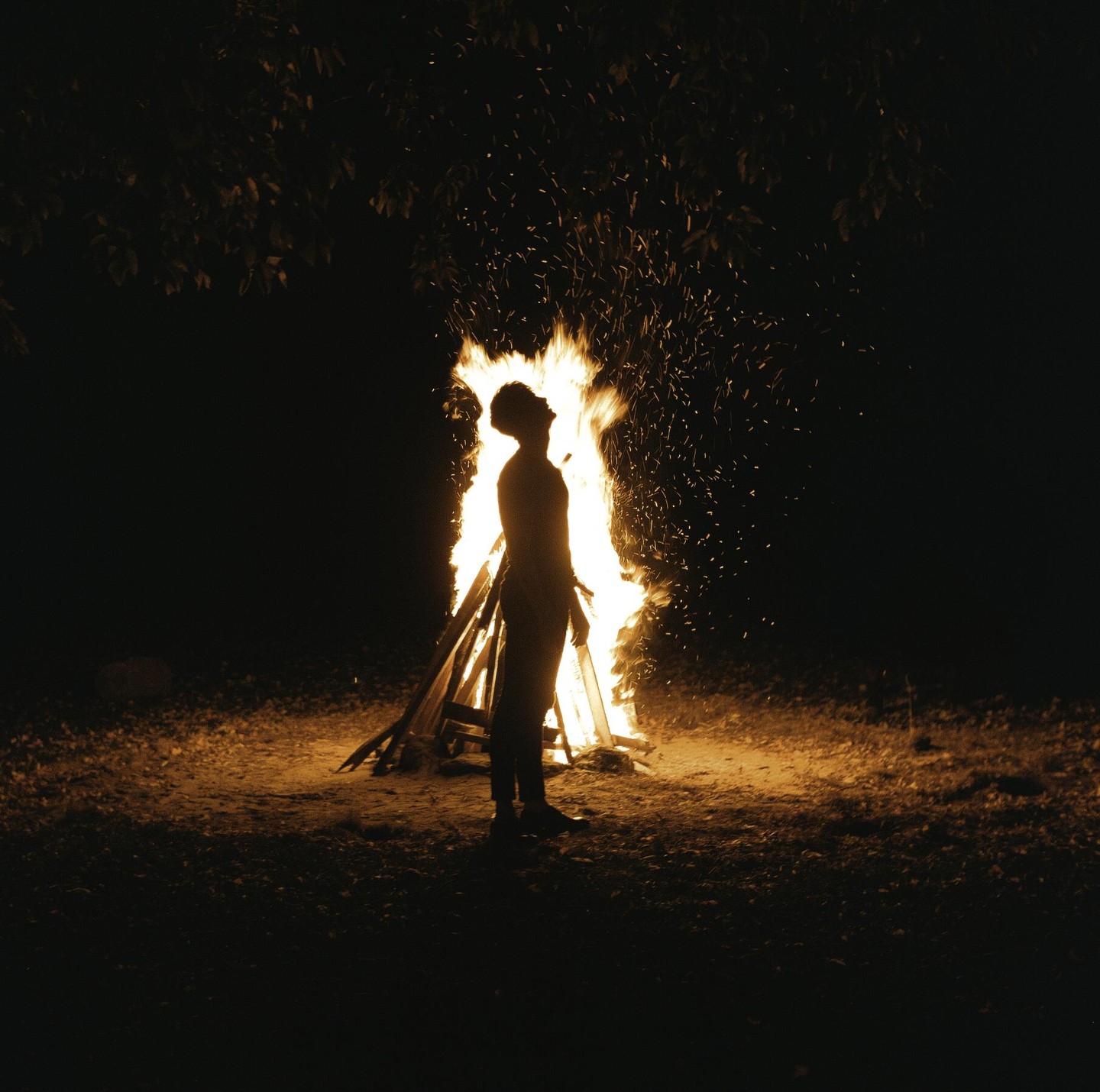 A man stands silhouetted against a large bonfire at night.