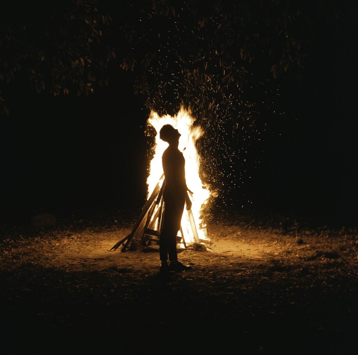 A man stands silhouetted against a large bonfire at night.