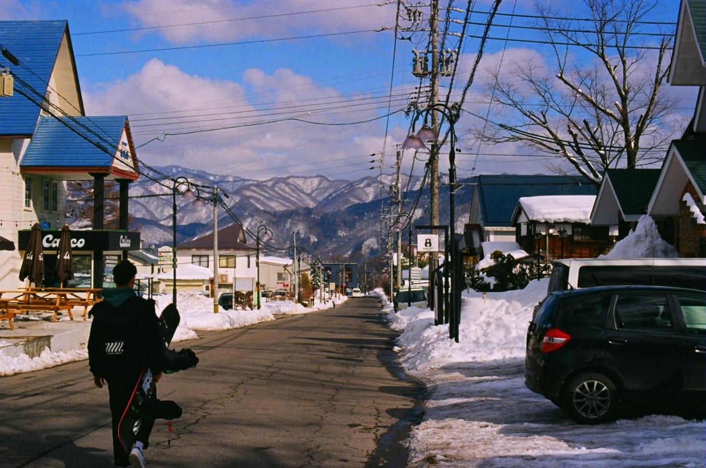 A man walks down a snowy street carrying a snowboard with mountains in the background.