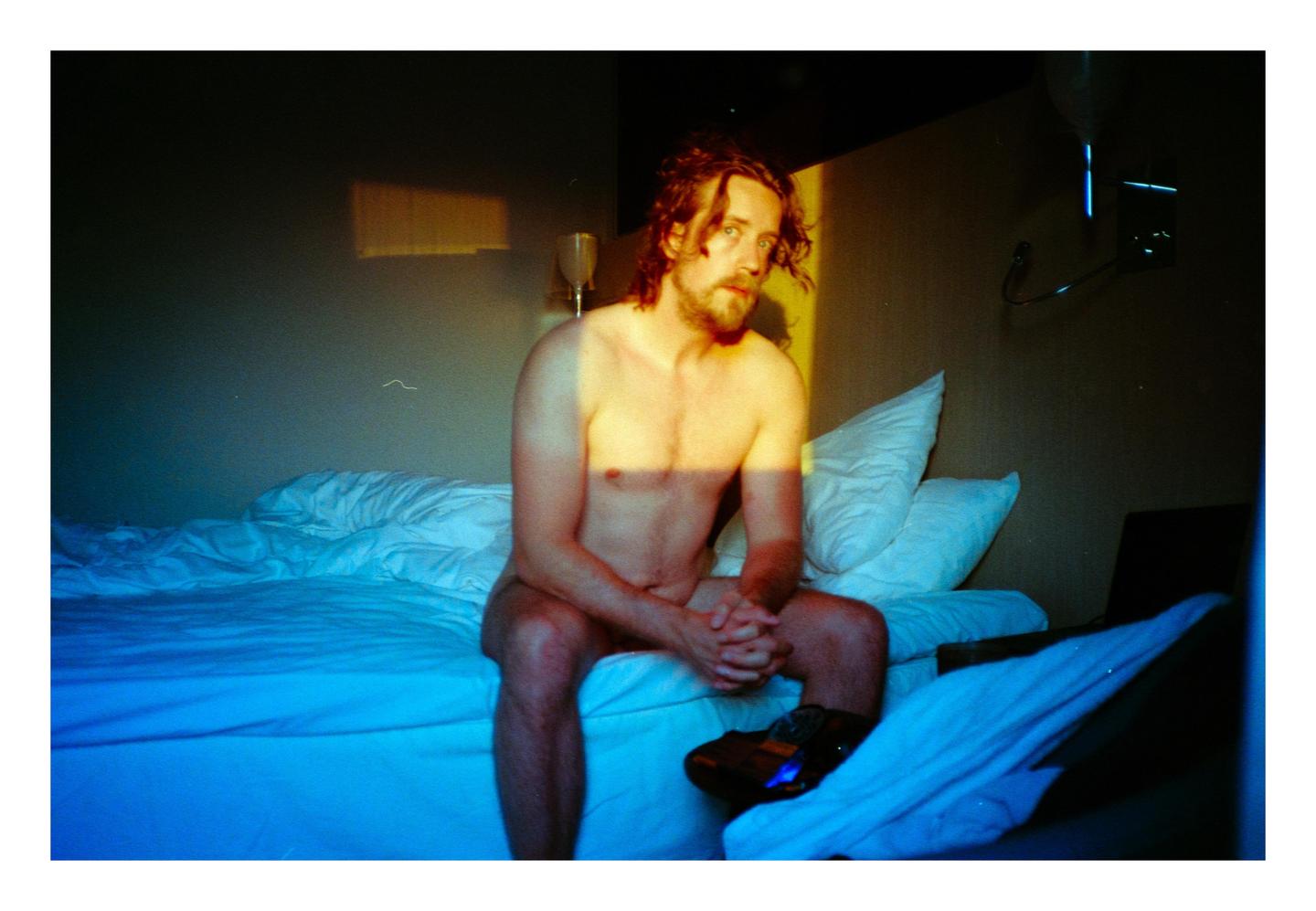 A shirtless man with long hair and a beard sits on a bed in a hotel room, illuminated by golden light.
