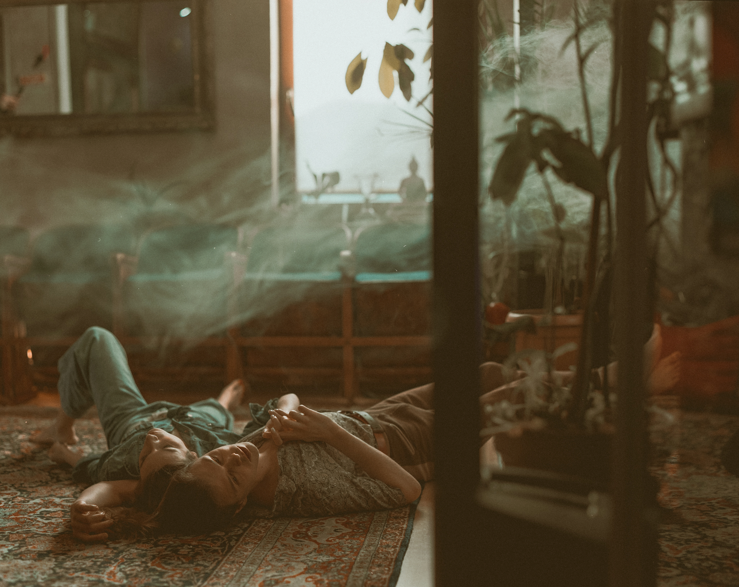 A couple lies on a patterned rug in a room filled with plants and soft light.