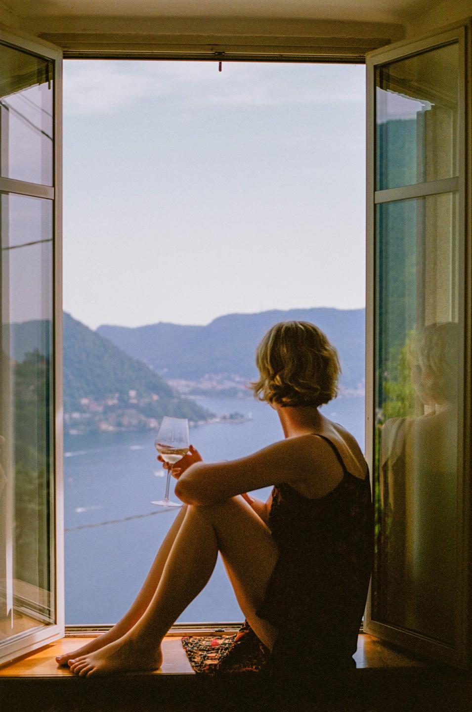 A woman sits in a window frame, holding a glass of wine and looking out at Lake Como.