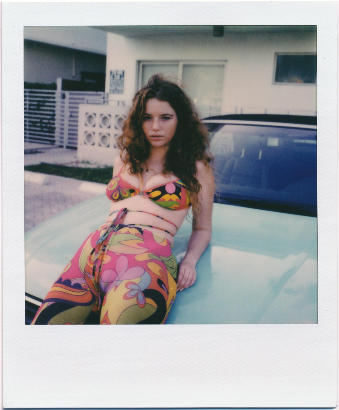 A woman in a colorful patterned outfit leans on the hood of a light blue car.
