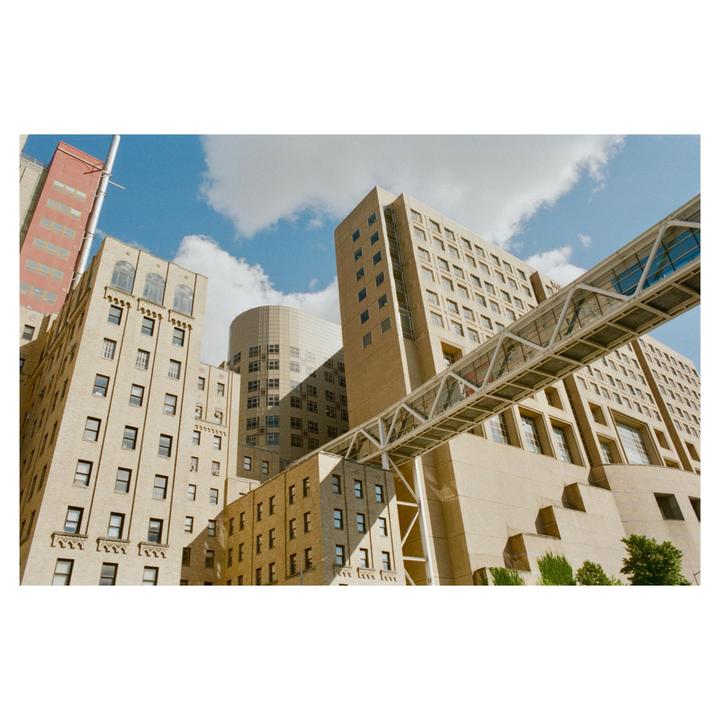 A low-angle view shows several tall buildings with a pedestrian bridge connecting two of them under a blue sky with clouds.