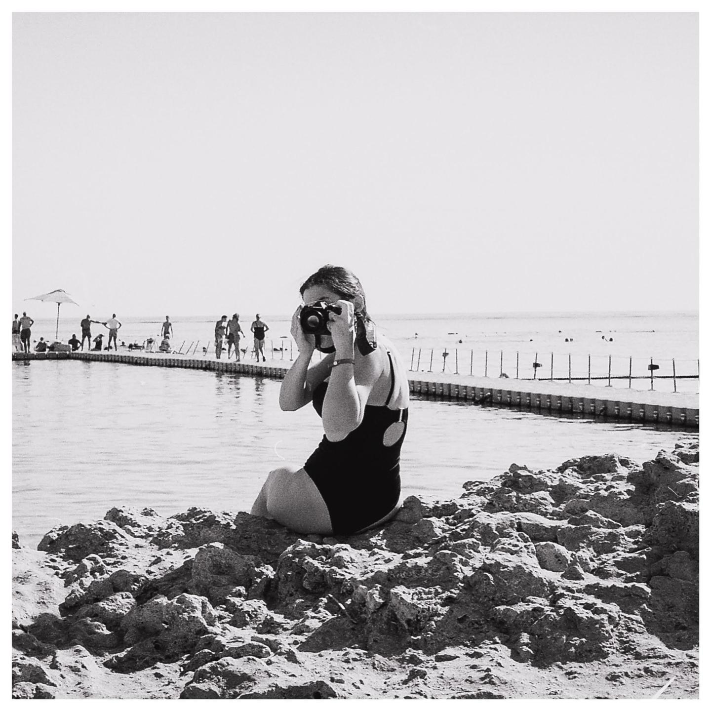 A woman in a swimsuit sits on rocks by the water and holds a camera to her face.