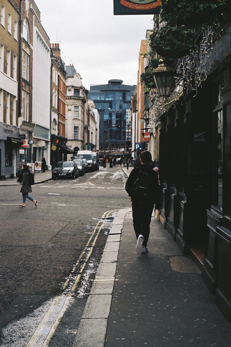 A person walks down a narrow street lined with buildings and parked cars, with another person crossing the street in the distance.