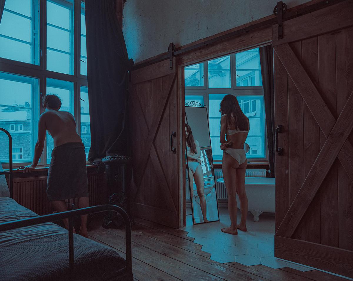 A man in a towel looks out a window while a woman in underwear stands in a doorway looking at her reflection in a mirror.