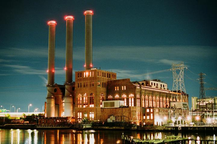 A large brick power plant with three tall smokestacks and glowing red tops stands beside a body of water at night.