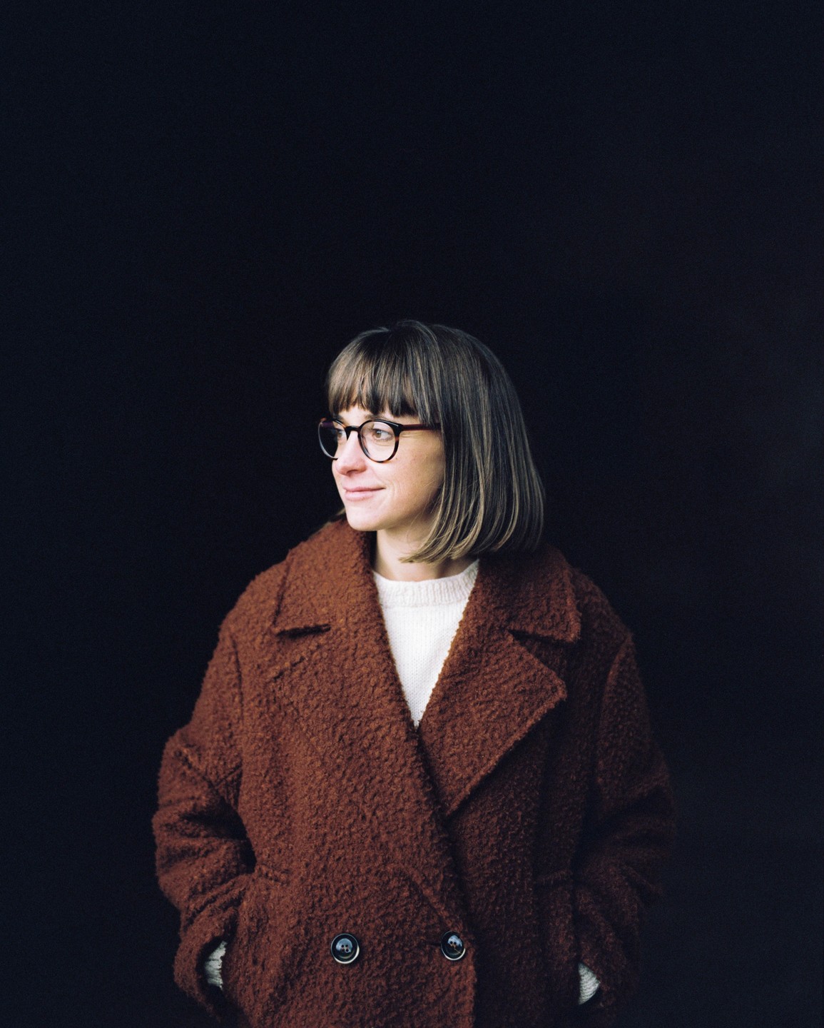 A woman wearing a brown coat and glasses looks to the left against a dark background.