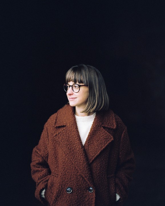 A woman wearing a brown coat and glasses looks to the left against a dark background.