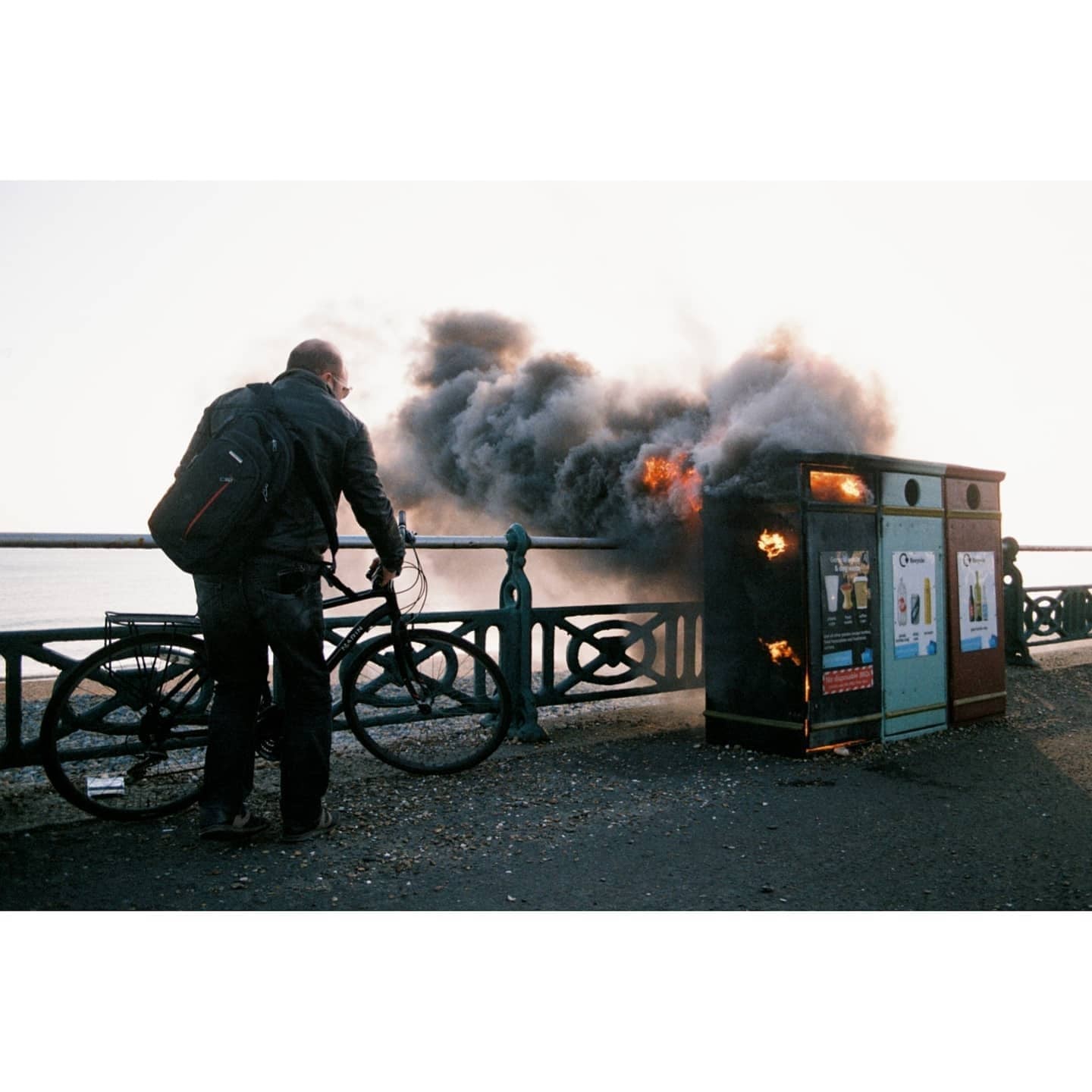 A man with a bicycle stands next to a burning trash can on a street.