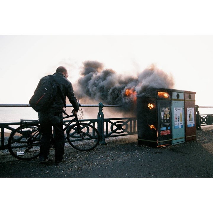 A man with a bicycle stands next to a burning trash can on a street.