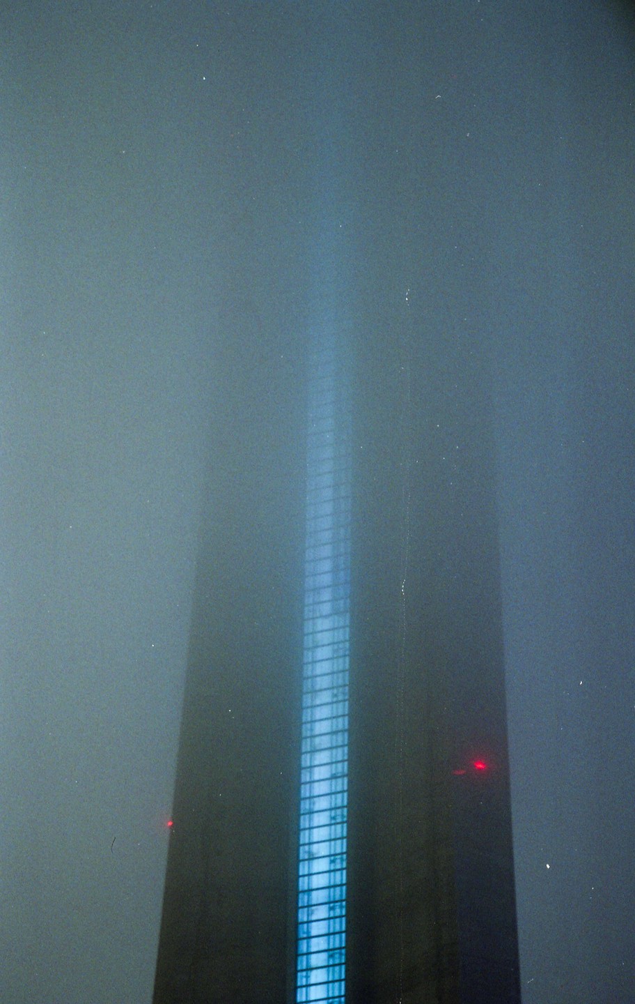 A tall building with a bright blue illuminated side rises into a foggy night sky.