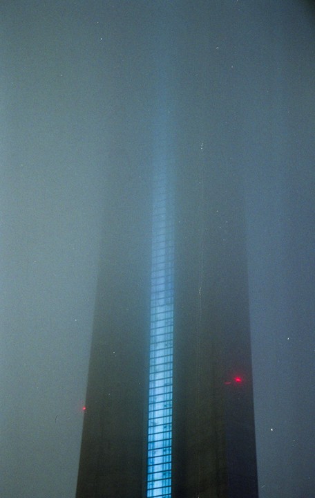 A tall building with a bright blue illuminated side rises into a foggy night sky.