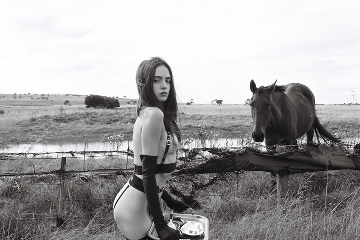 A woman in lingerie holds a tray with a teacup and saucer while standing in a field with a horse behind a fence.