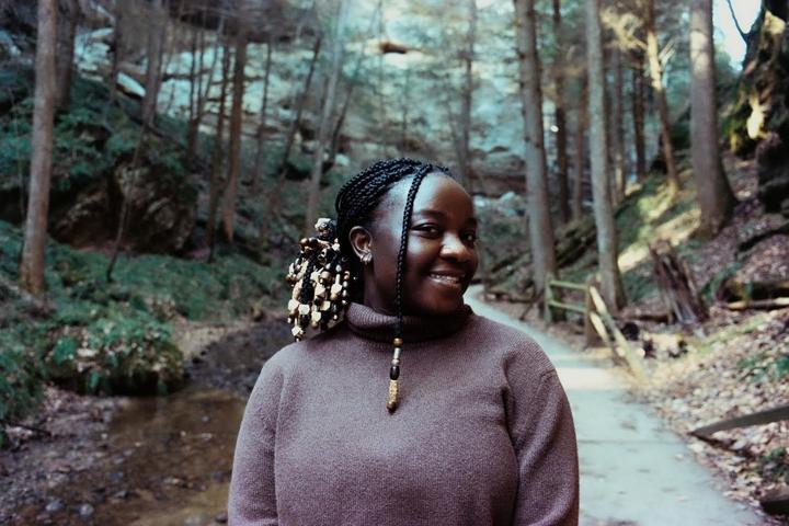 A woman with braided hair and beads smiles while standing in a forest with a path and stream.