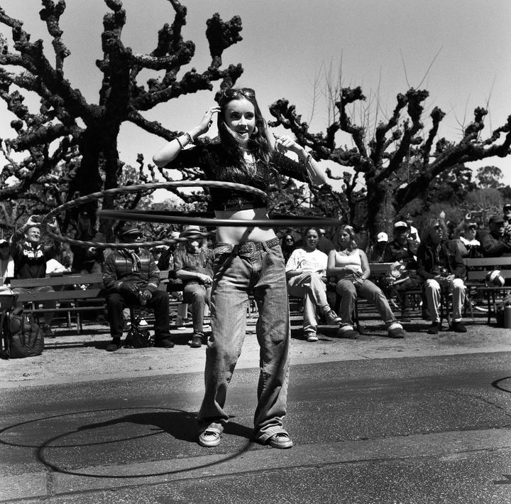 A woman hula hoops in front of a crowd of people sitting on benches under trees.