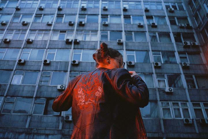 A person with a top bun and glasses stands with their back to the viewer, wearing a jacket with red embroidery, in front of a building with many windows and air conditioning units.
