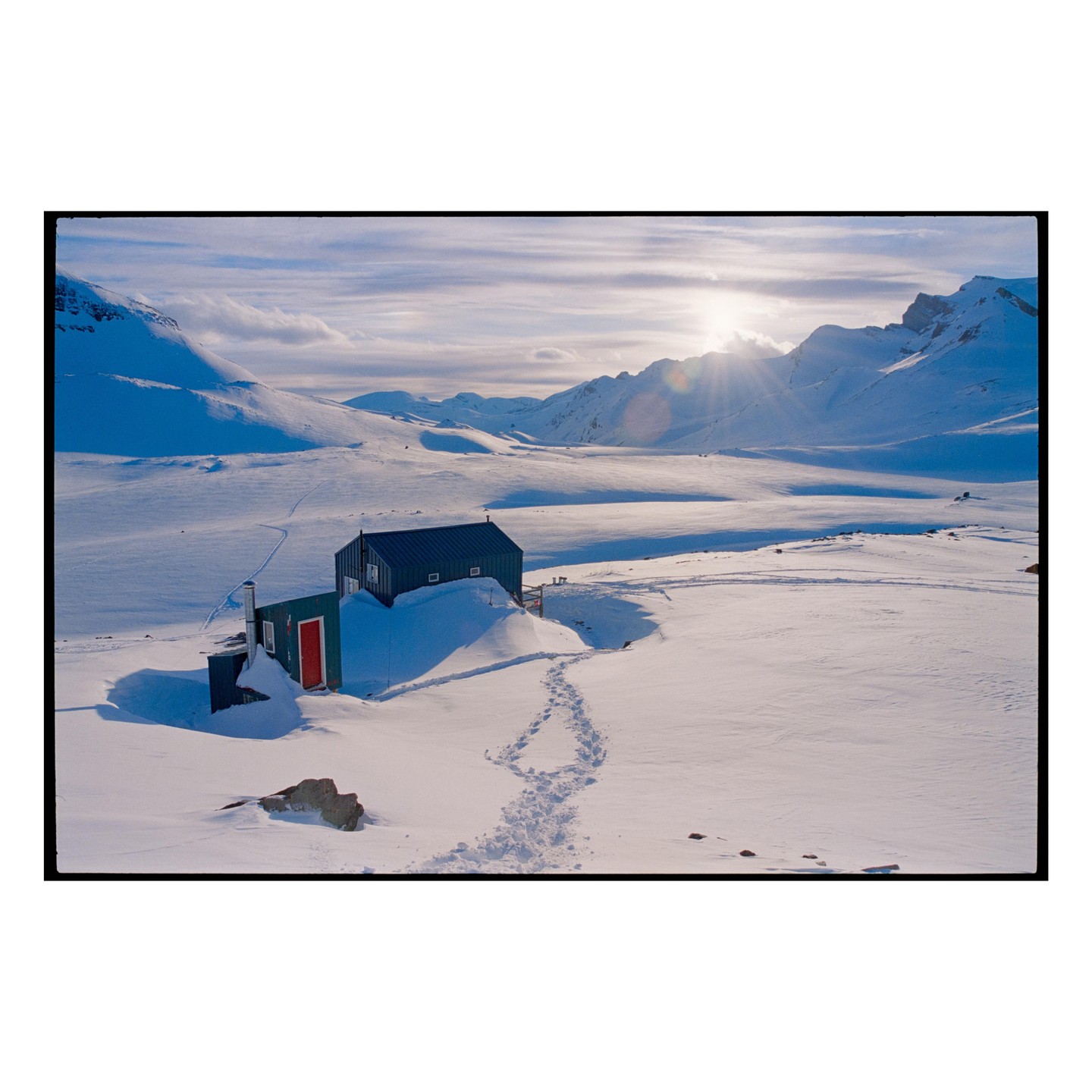 A small cabin with a red door is nestled in a vast, snow-covered mountain landscape under a bright, sunlit sky.