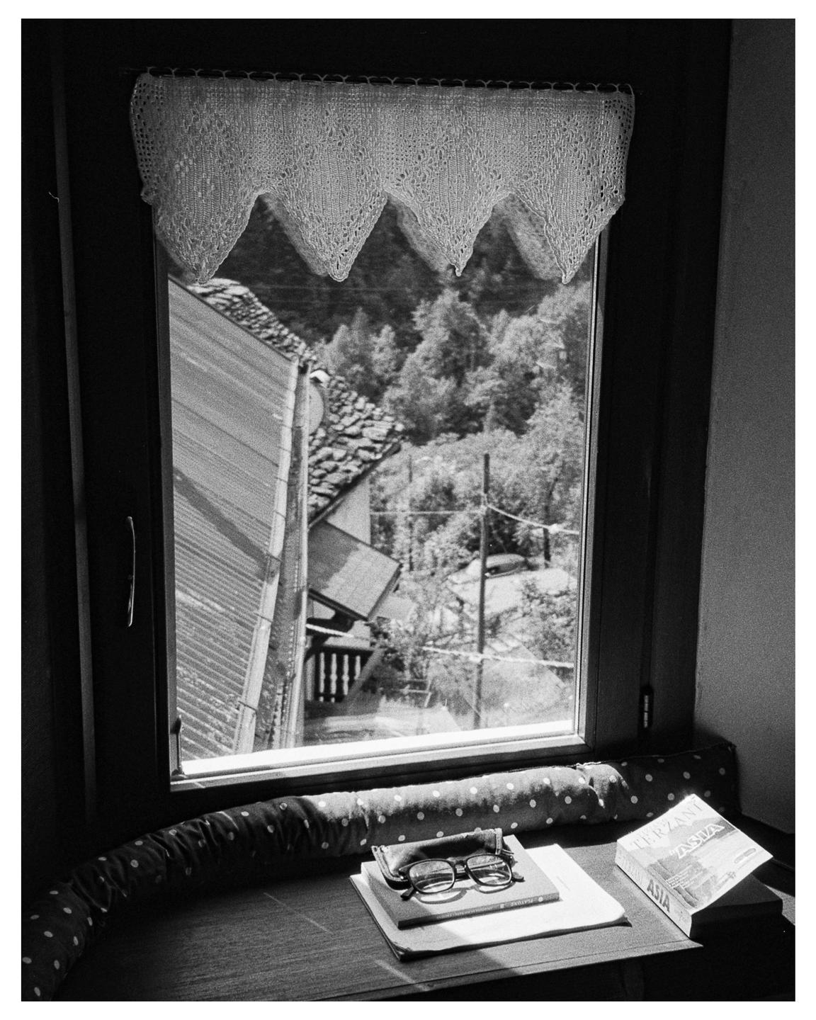 A pair of eyeglasses rests on a book next to a window with a view of a village and trees.