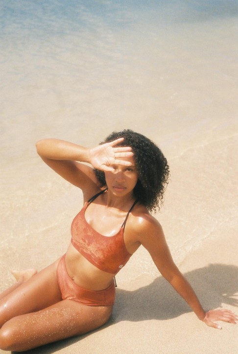 A woman in a bikini sits on a sandy beach with her hand shielding her eyes from the sun.