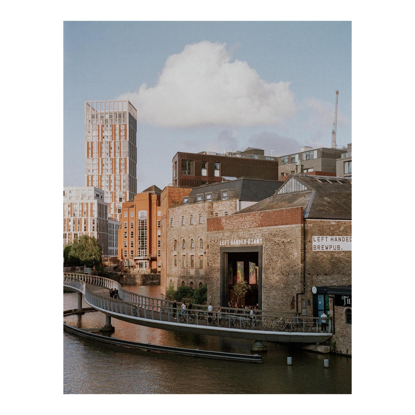 A modern pedestrian bridge crosses a river in a city with a mix of old and new buildings under a partly cloudy sky.