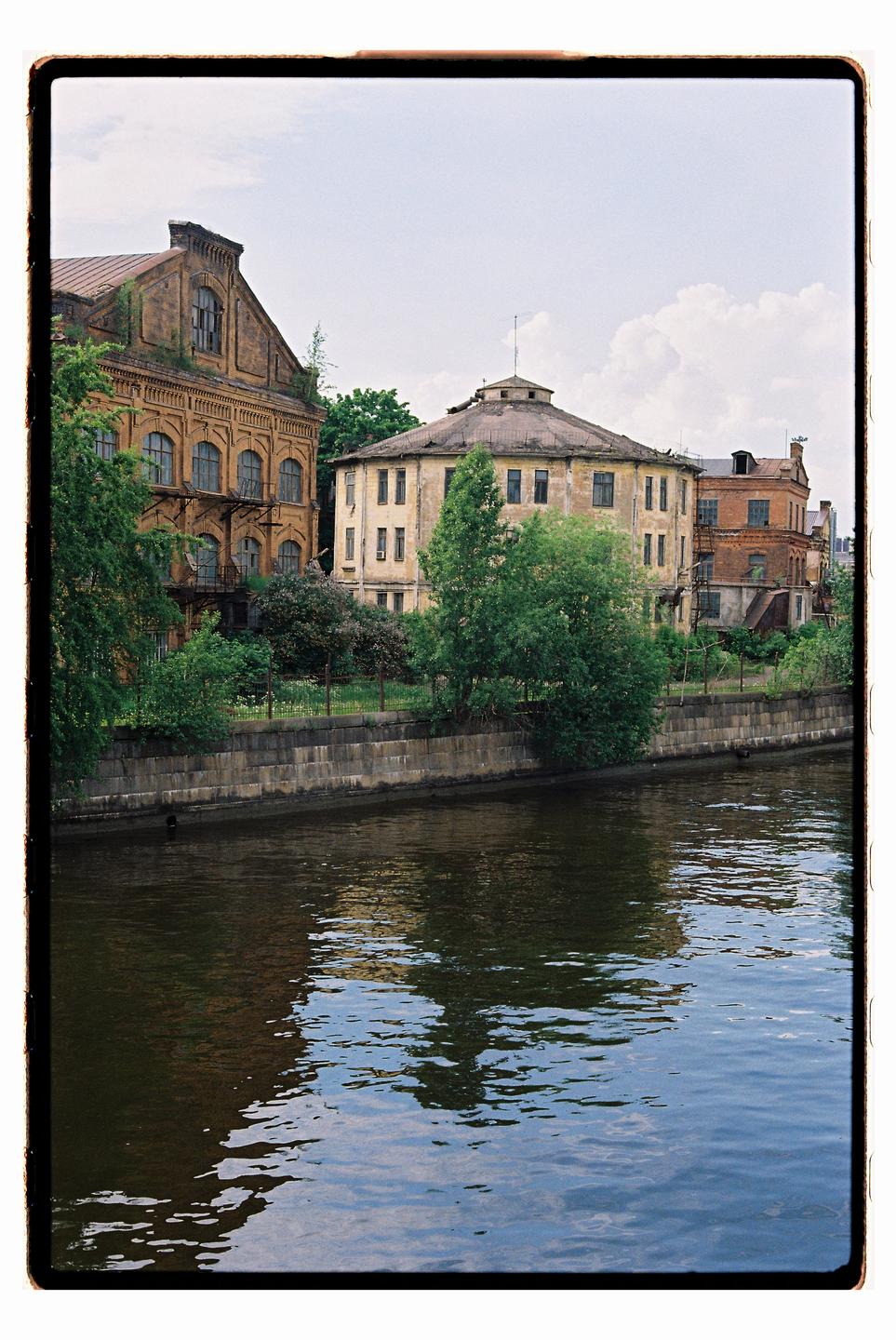 A river flows past old buildings and green trees under a cloudy sky.