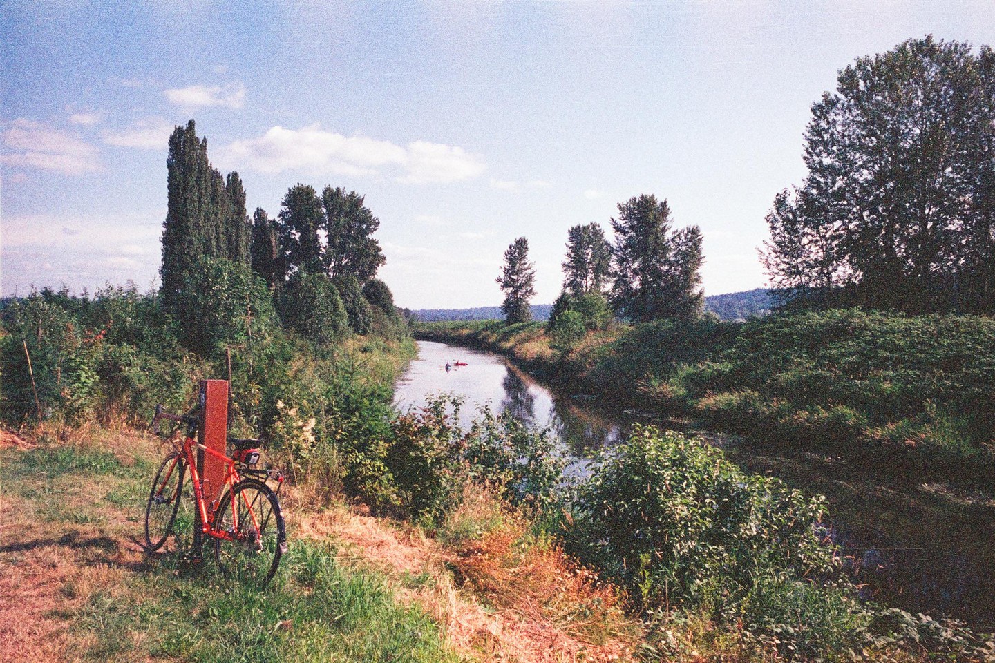 A red bicycle is parked on a grassy bank next to a narrow river where two people are kayaking.