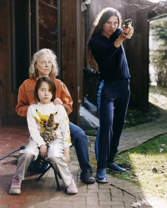 A woman aims a handgun while a child sits on a chair in front of an older woman.
