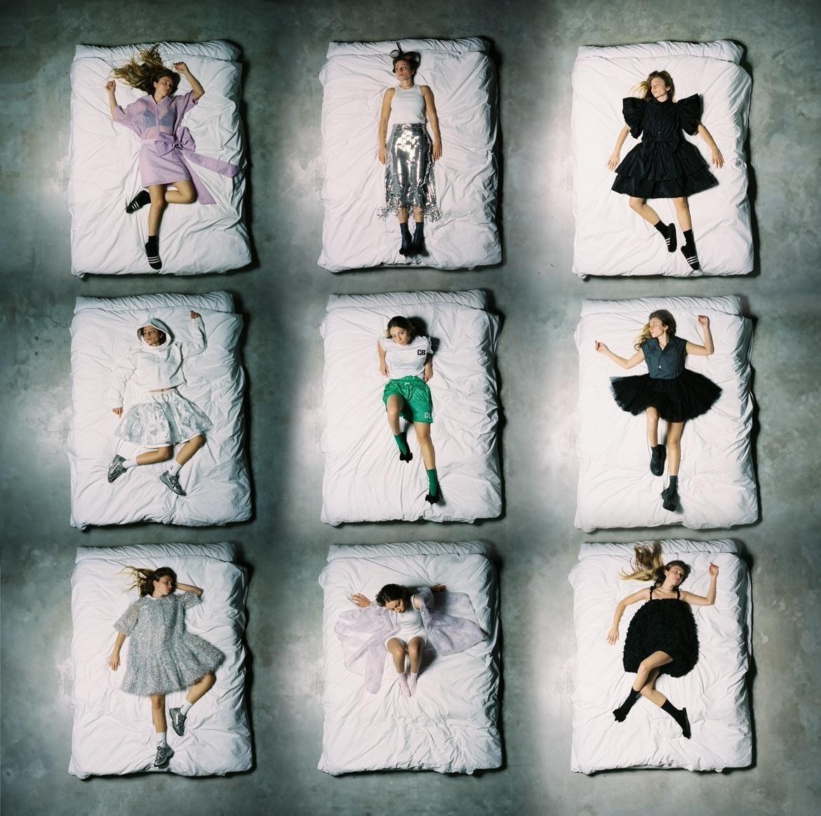 Nine women lie on nine separate beds arranged in a grid pattern on a concrete floor.