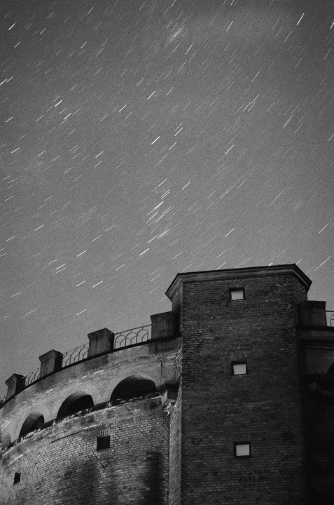 A brick building with arched windows and a railing on the roof is visible under a night sky filled with star trails.