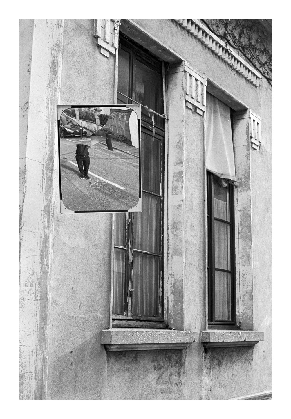 A mirror reflecting a person's legs and a car is attached to the side of a building with two windows.