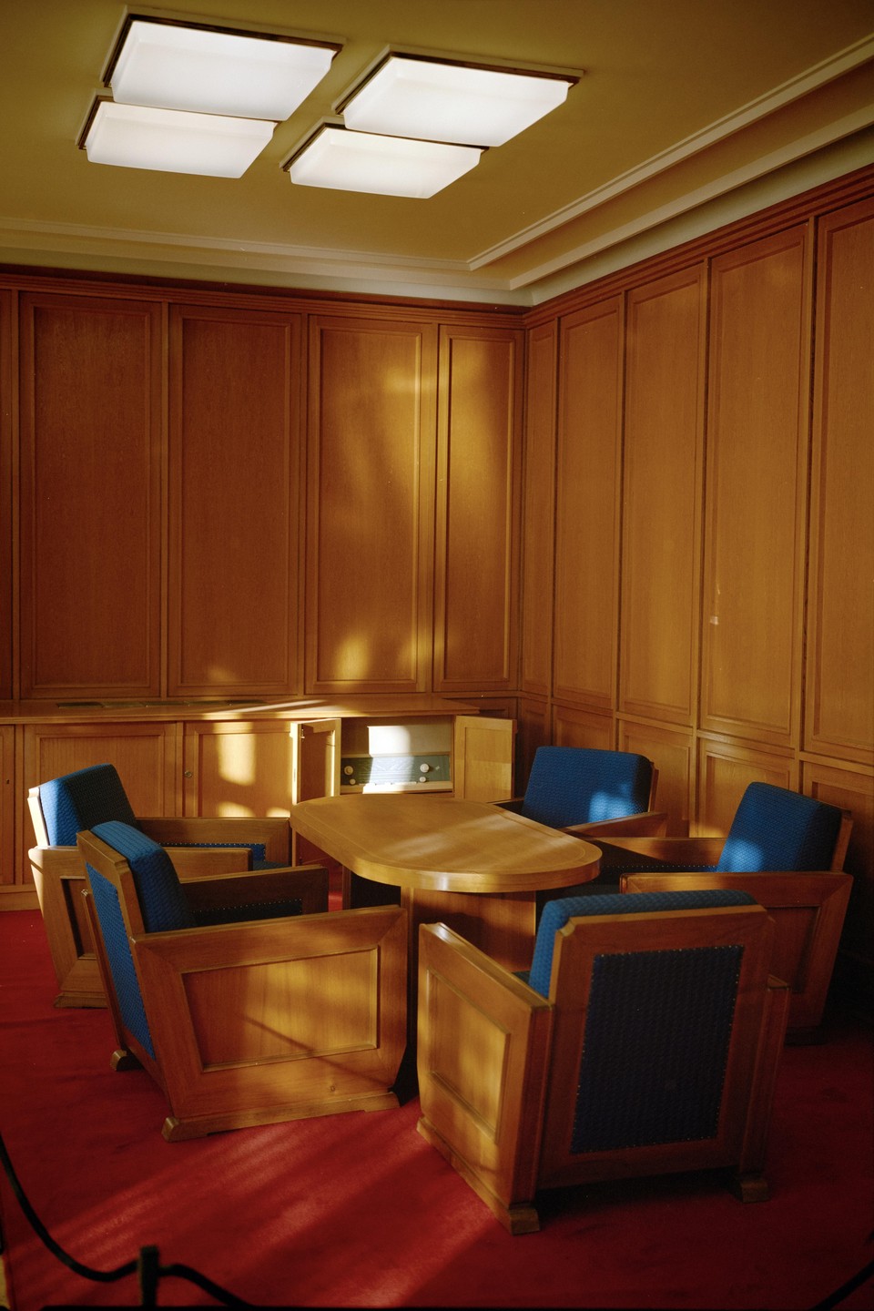 A room with wood paneling, a red carpet, a wooden table, and several armchairs with blue upholstery is illuminated by overhead lights and sunlight streaming through a window.