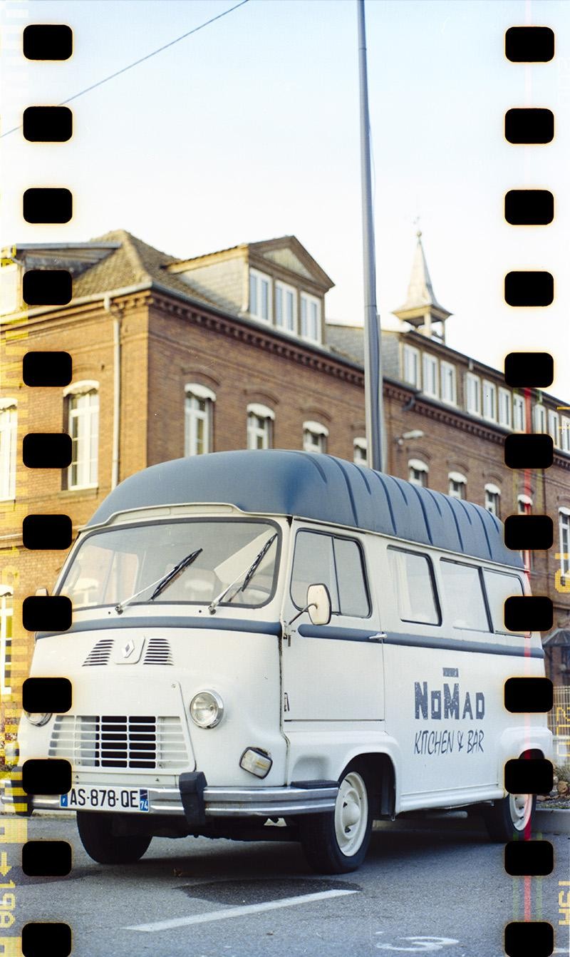 A white Nomad Kitchen & Bar van is parked on a street in front of a brick building with a steeple.