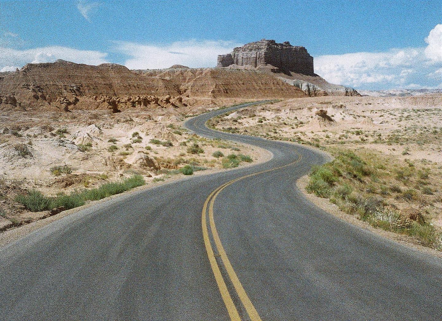 A winding road with double yellow lines leads through a desert landscape towards a large mesa under a blue sky with clouds.
