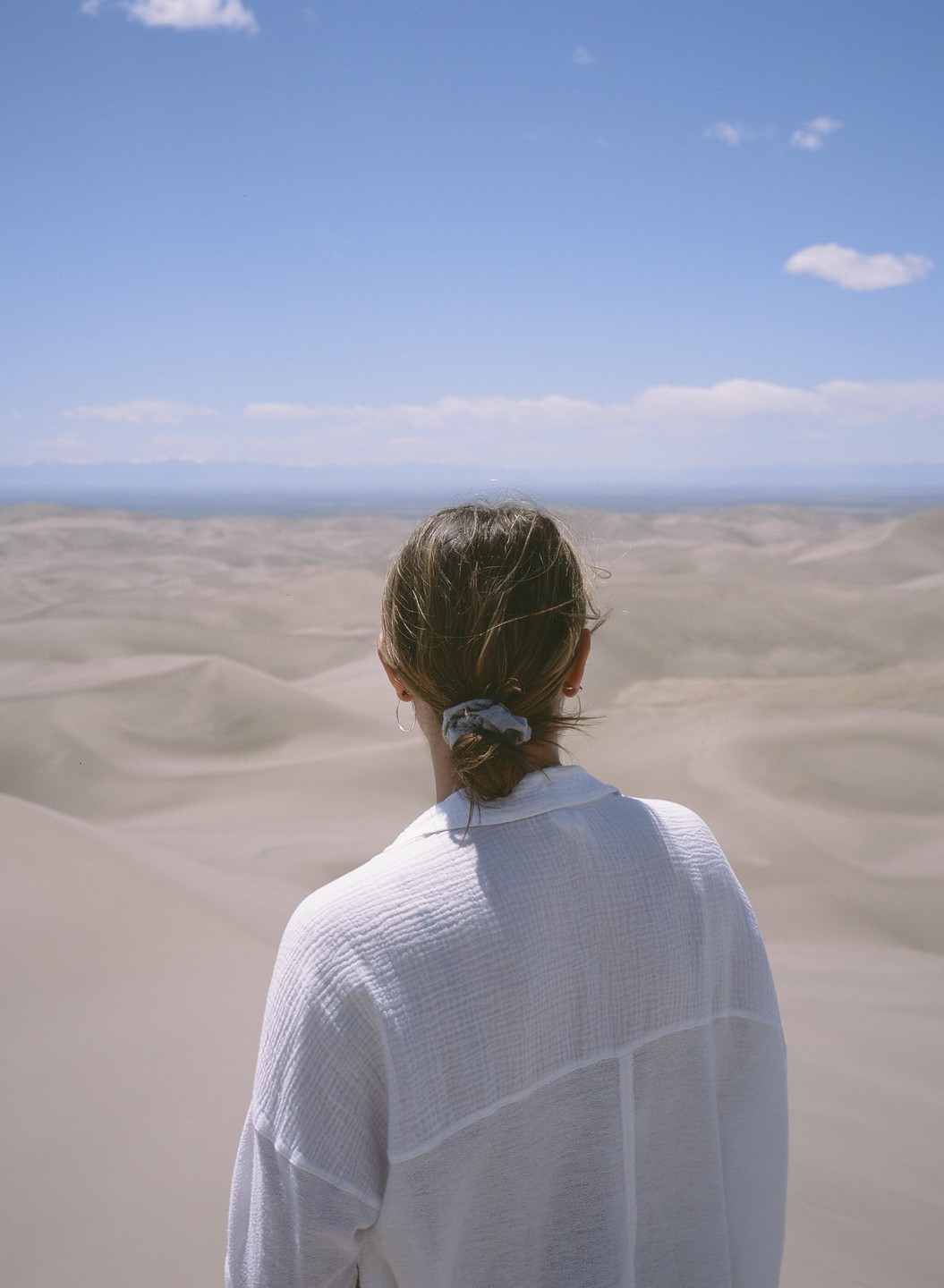A woman in a white shirt looks out over a vast desert landscape with sand dunes under a blue sky.